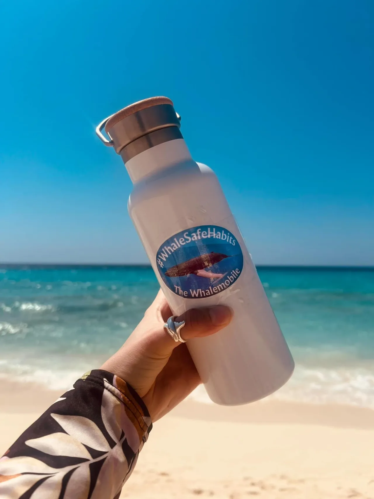 A hand holding a bottle with a WhaleSafeHabits sticker in front of the ocean